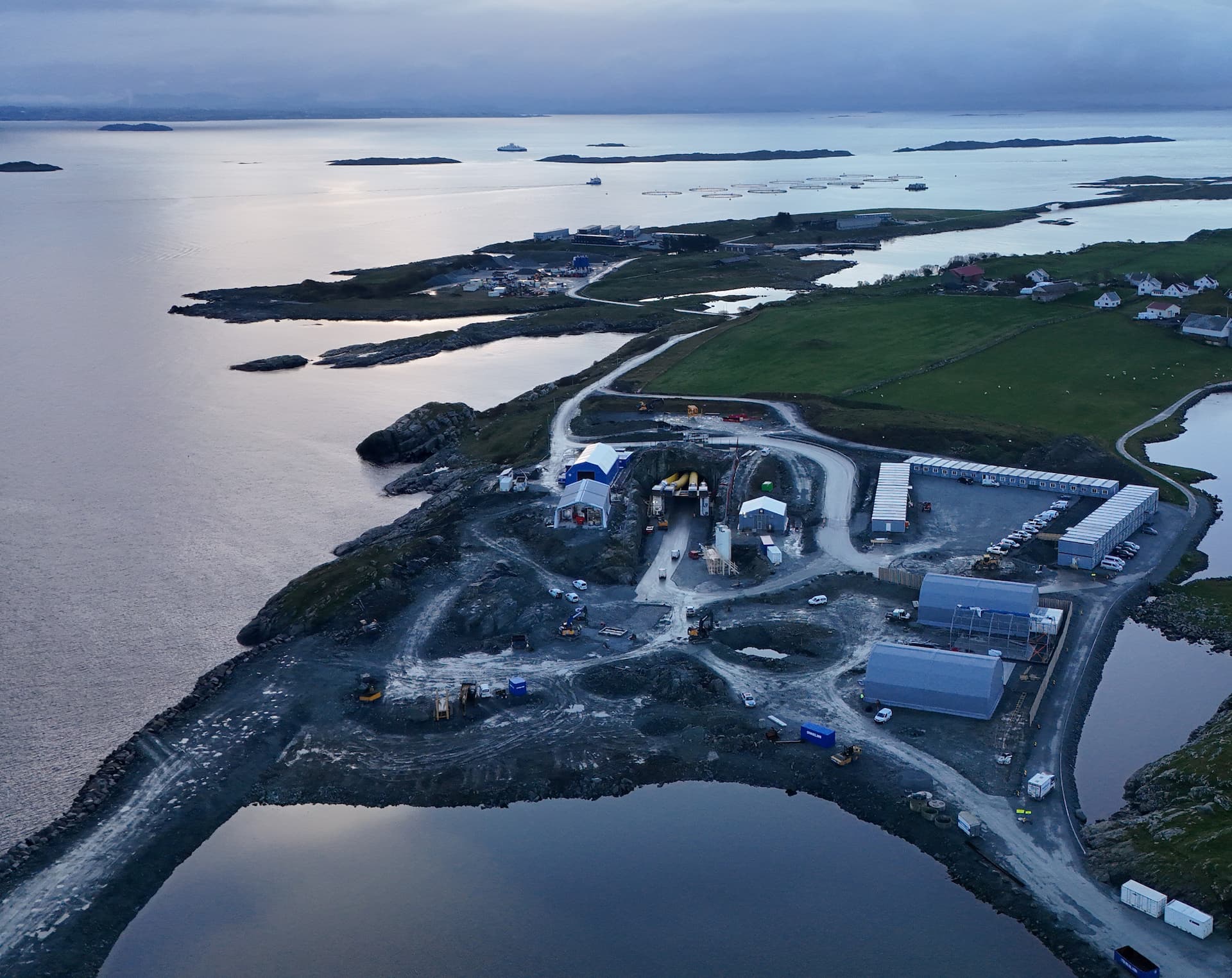 Anleggsområde som ligger like ved havet. Midt i ligger tunnelåningen som for tunnelen fra Kvitsøy til Rogfast. Bondegårder og lakseoppdrettsanlegg i bakgrunnen.